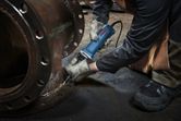A person wearing safety equipment uses a straight grinder on a large metal flange.