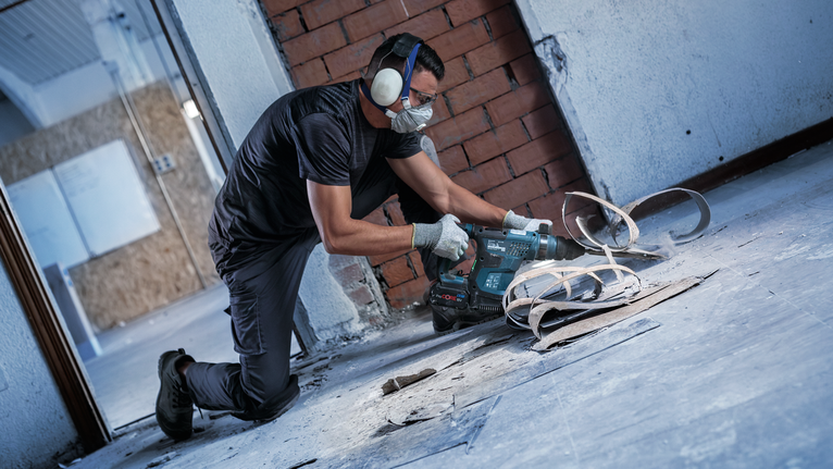 Person wearing safety equipment uses a rotary hammer to break concrete on a construction site.