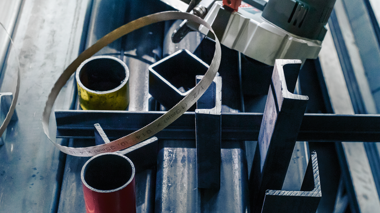 PRO CB281418 Band Saw Blade, 733 mm, 2 pcs Band saw poised above assorted metal pipes and bars on a workbench.