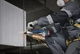 A person wearing safety equipment grinds a metal beam, creating sparks.