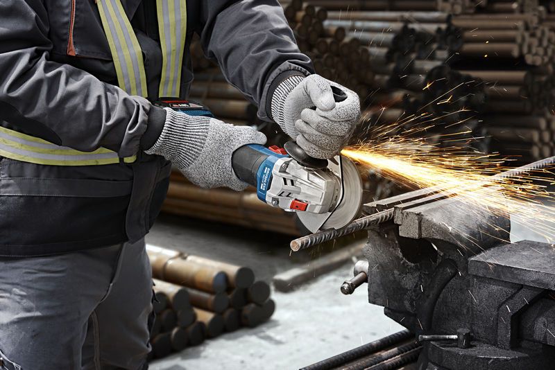 A person wearing safety equipment uses an angle grinder to cut a metal rod, producing sparks.