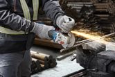A person wearing safety equipment cuts metal rebar with an angle grinder, sparks flying.