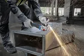A person wearing safety equipment grinds a metal duct, producing sparks.