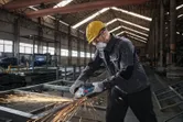 A person wearing safety equipment grinds metal, producing bright sparks in a factory.