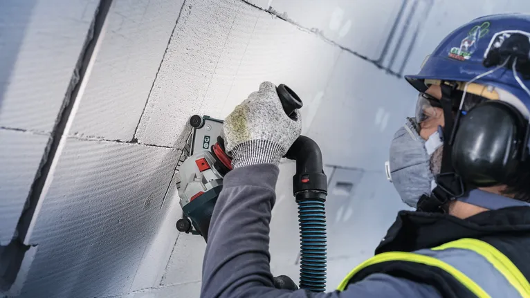 Person wearing safety equipment grinds into a concrete wall using a power tool.