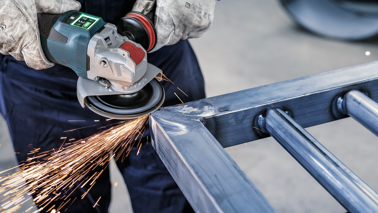 PRO Backing Pad Hard, 125 mm, X-Lock Person wearing safety equipment grinds a metal beam with an angle grinder, creating sparks.