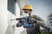 Person wearing safety equipment drills into a wall with a cordless hammer drill.