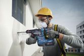 A person wearing safety equipment drills into an exterior wall with a power tool.
