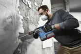 A person wearing safety equipment uses a rotary hammer to remove tiles from a wall.