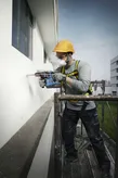 Worker wearing safety equipment drills into a building facade while standing on scaffolding.
