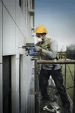 A person wearing safety equipment drills into an exterior building wall on scaffolding.