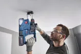 A person wearing safety equipment drills into a ceiling with a rotary hammer.