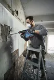 A person wearing safety equipment removes wall tiles using a rotary hammer.
