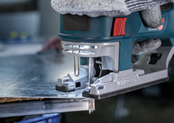 Person wearing safety equipment uses a jigsaw to cut a metal sheet.