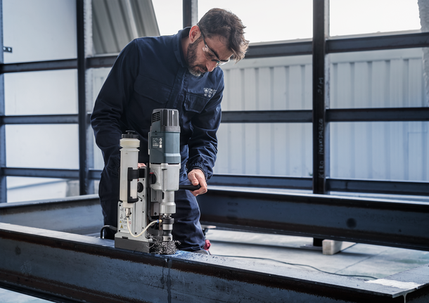 Person wearing safety equipment operates a magnetic drill on a steel beam.