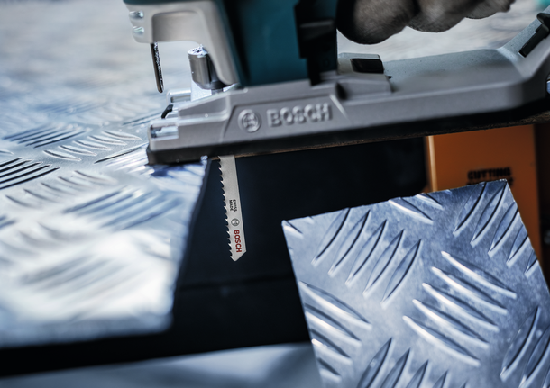 Person cuts textured metal sheet with a jigsaw, wearing safety equipment.