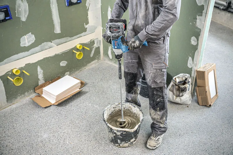 A person wearing safety equipment mixes material in a bucket using a stirring mechanism.