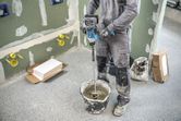 A person wearing safety equipment mixes material in a bucket using a stirring mechanism.