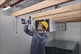 A person wearing safety equipment uses a power drill to fasten metal supports overhead.