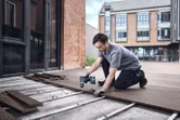 A person wearing safety equipment uses a cordless drill to install decking boards outside.