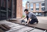 A person wearing safety equipment uses a cordless drill to install decking boards outside.