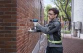 A person wearing safety equipment drills into a brick wall outdoors.