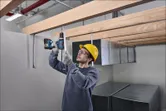 A person wearing safety equipment drills into a metal rod beneath wooden beams.