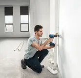 A person drills into a wall beside plumbing pipes in a partially finished room.
