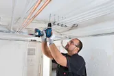 A person wearing safety equipment drills into a ceiling to install electrical conduit.