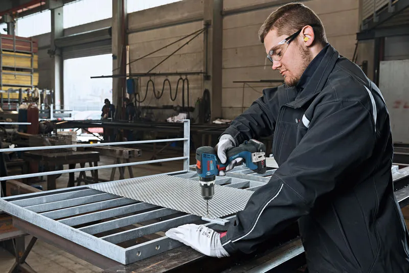 A person wearing safety equipment drills metal mesh on a workbench in a factory.