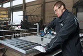 A person wearing safety equipment drills metal mesh on a workbench in a factory.
