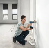A person drills into a wall to install piping in a bright, unfinished room.