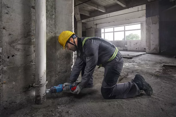 A person wearing safety equipment uses a reciprocating saw to cut a pipe near a concrete wall.