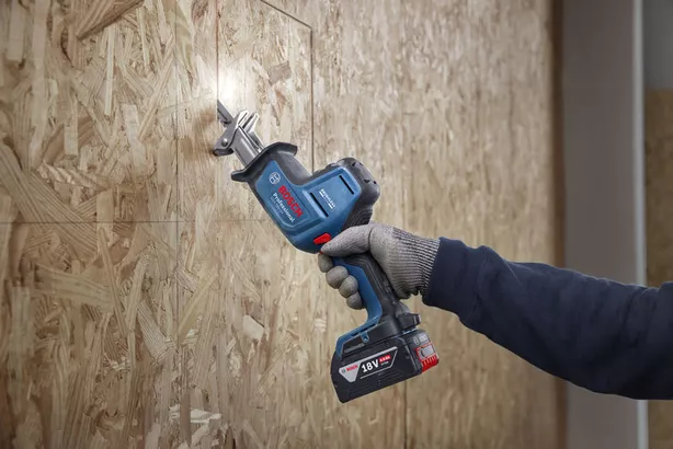 Person wearing safety equipment cuts wood panel using a cordless reciprocating saw.