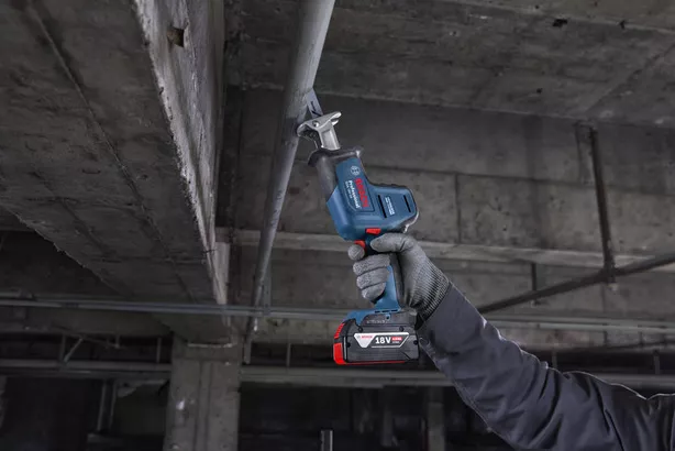 A person wearing safety equipment cuts a metal pipe overhead with a reciprocating saw.