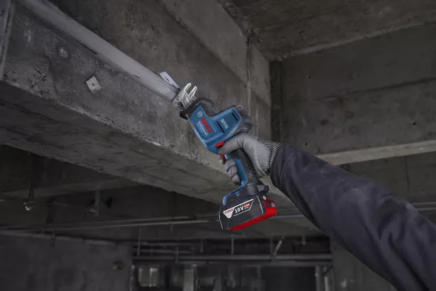 A person wearing safety equipment uses a reciprocating saw to cut metal overhead.