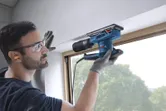 A person wearing safety equipment sands a window frame with an orbital sander.