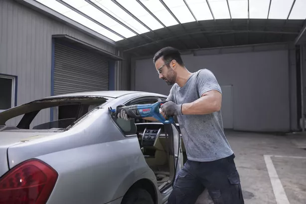 Person wearing safety equipment uses a reciprocating saw to cut a car’s rear window frame.