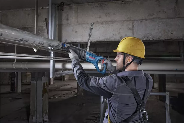 A person wearing safety equipment cuts a metal pipe with a reciprocating saw.