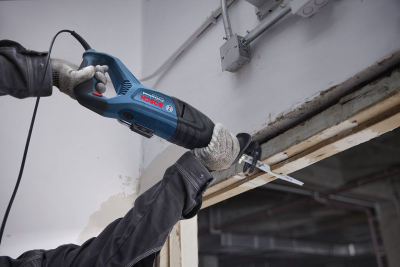 A person wearing safety equipment operates a reciprocating saw to cut a wooden beam.
