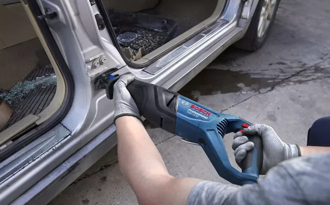 A person wearing safety equipment uses a reciprocating saw to cut metal on a car door frame.
