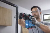 A person wearing safety equipment drills into a metal frame using a cordless rotary hammer.