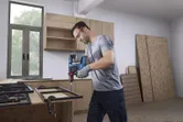 A person wearing safety equipment drills metal frames on a wooden workbench.