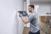 A person wearing safety equipment drills into a marked wall with a cordless rotary hammer.