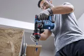 A person wearing safety equipment drills into wood with a cordless rotary hammer.