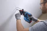 A person wearing safety equipment drills a hole into a marked wall with a cordless rotary hammer.