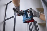 A person wearing safety equipment drills a hole in a metal frame with a cordless rotary hammer.