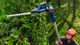 A person wearing safety equipment trims tall hedges with a cordless hedgecutter.