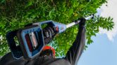 A person wearing safety equipment trims high branches with a battery-powered hedgecutter.