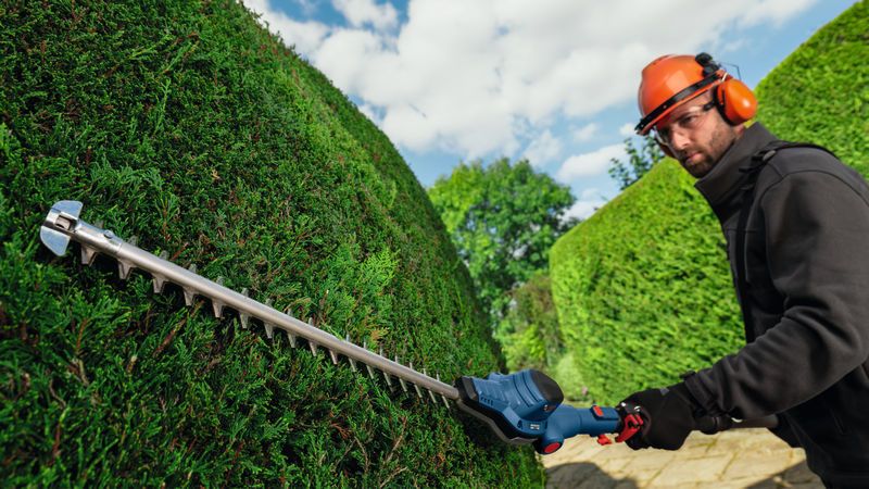 GHE 18V-50 TP A person wearing safety equipment trims hedges with a cordless telescopic hedgecutter.